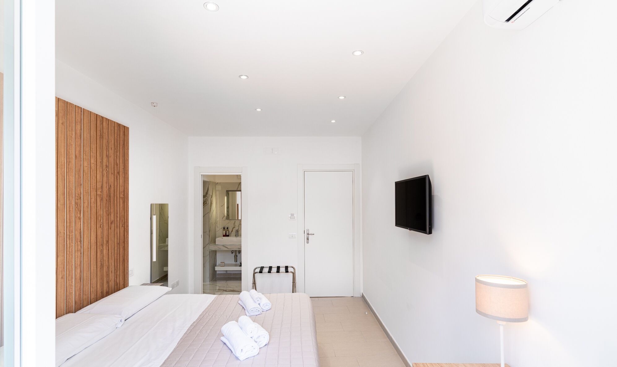 Habitación de hotel moderna con una cama, un televisor montado en la pared, una lámpara de noche y un panel de madera en la pared izquierda.