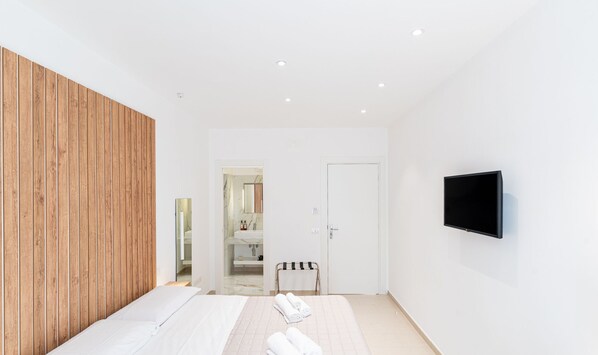 A modern bedroom with a wooden accent wall, a bed with white linens, a TV mounted on the wall, and a bathroom visible through an open door. - Olivia's Home By DomusExtra (Naples)