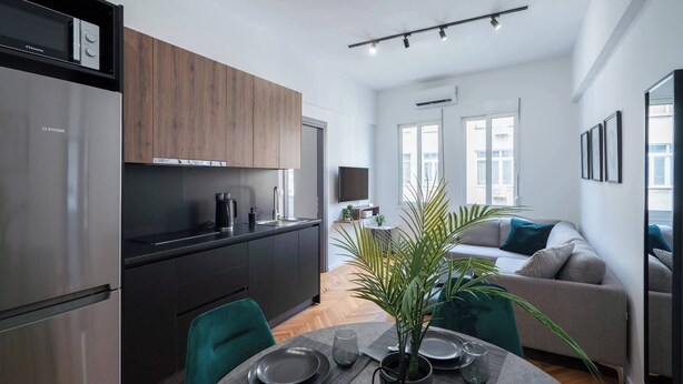 A modern kitchen with a built-in oven, a dining area with a table set for two, and a living area with a sofa and a plant.