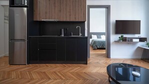 A modern kitchen with a built-in microwave, a stainless steel refrigerator, and a wooden cabinet. There is a black countertop with a sink and a coffee machine. The room has a wooden herringbone floor and a black glass table.