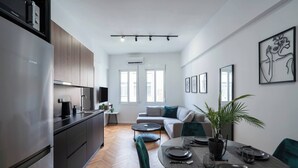 A modern kitchen with a stainless steel refrigerator, wooden cabinets, and a herringbone wood floor. There is a dining area with a round table, green upholstered chairs, and a potted plant. The room has large windows and a view of the outside.