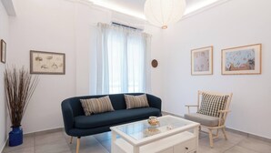 A living room with a dark blue sofa, a white coffee table, a wooden chair, and a large window with sheer curtains. - Bright 2BR Apartment By Metro Near Acropolis (Athens)