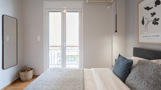 Een slaapkamer met een bed, een raam, een airconditioner, een ingelijst beeld en een mand op de vloer.