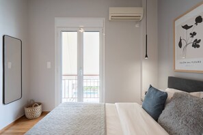 A bedroom with a bed, a window, an air conditioner, a framed picture, and a basket on the floor.