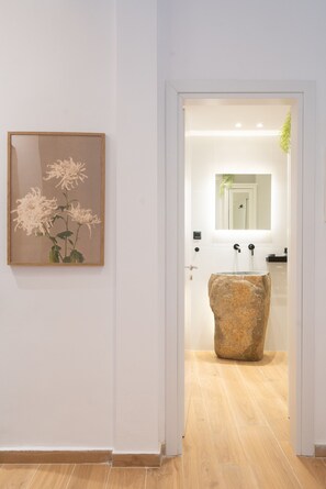 A hallway with a framed artwork of flowers, a stone sink, and a mirror in a bathroom. - 1BR Stylish Apt 20 Minutes from Acropolis (Athens)