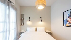 Une chambre à coucher comprenant un lit, deux tables de chevet, une étagère murale agrémentée d’objets décoratifs, un luminaire suspendu et un tableau encadré accroché au mur.