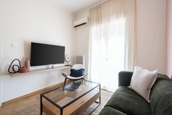 A modern living room with a flat-screen TV, a glass coffee table, a green sofa, and a chair with a cushion.