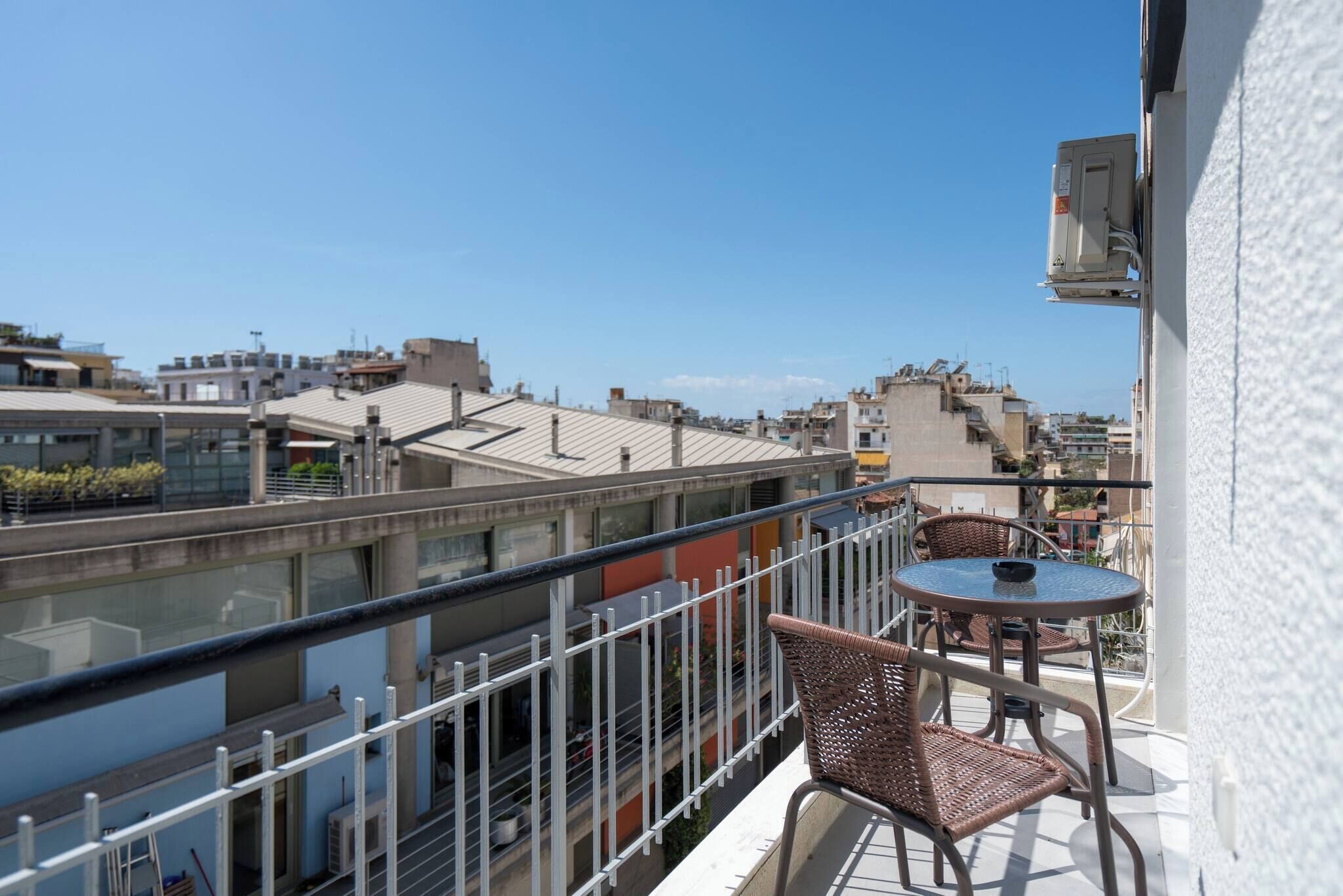 A balcony with wicker chairs and a small table, overlooking a cityscape with various buildings.