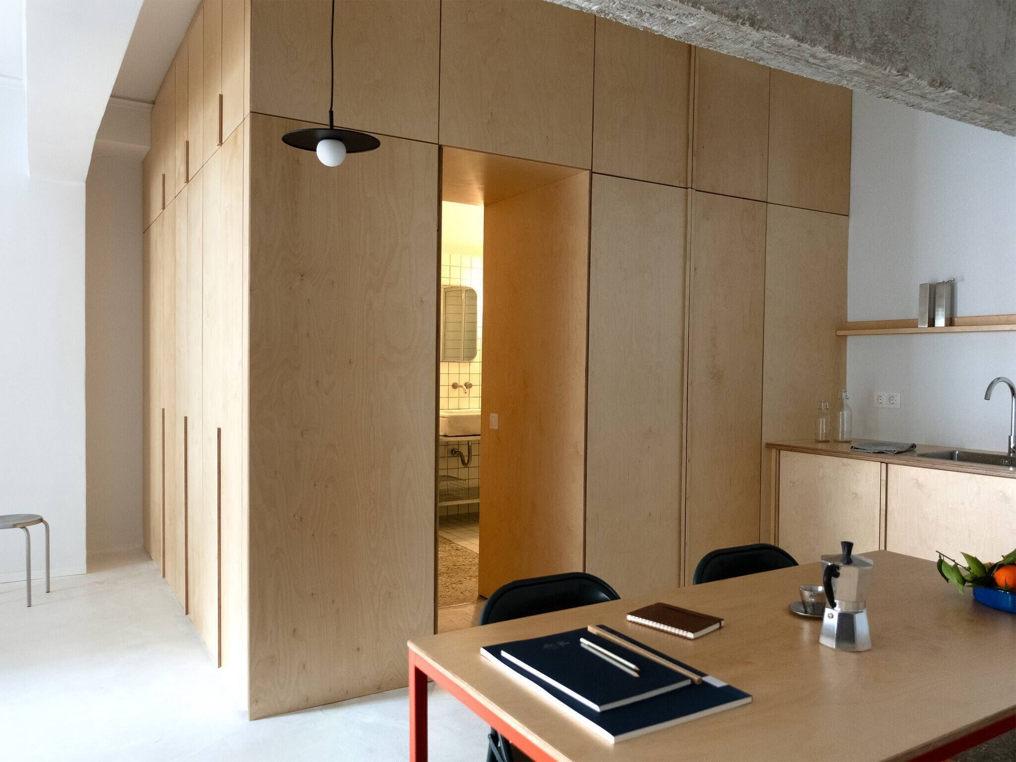 A modern kitchen with wooden cabinetry, a dining table, chairs, a coffee maker, and a view into the adjacent bathroom.