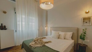 Un dormitorio ordenado con cama, mesita de noche, lámpara, radiador y ventana con cortinas.