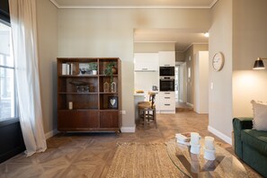 A modern living room with a wooden bookshelf, a green sofa, a glass coffee table with candles, and a kitchen in the background.
