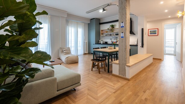 A modern living room with a sofa, dining table, and chairs. There is a kitchen area with cabinets and a countertop.
