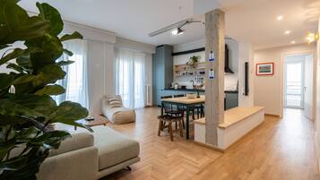 A modern living room with a sofa, dining table, and chairs. There is a kitchen area with cabinets and a countertop.