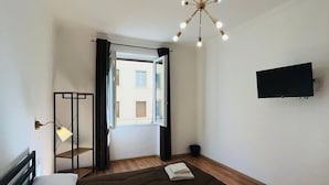 A modern bedroom with a large bed, a ceiling light fixture, a TV mounted on the wall, and a window with curtains. - Prime Rooms Della Loggia (Florence)