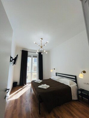 Camera d'albergo con un letto grande, un lampadario, una finestra con tende e vista sugli edifici esterni.