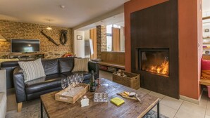 A cozy living room with a fireplace, a leather sofa, a coffee table with glasses and snacks, and a TV on a brick wall.