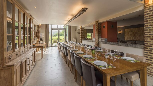 A long dining table set for a meal, with a kitchen area and a view of the outdoors.