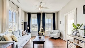 A spacious living room with a dark wooden coffee table, a grey sofa, and a white armchair. There is a ceiling fan, large windows with curtains, and a view of the outside. - The Happy Hut - walk to the beach (Galveston)