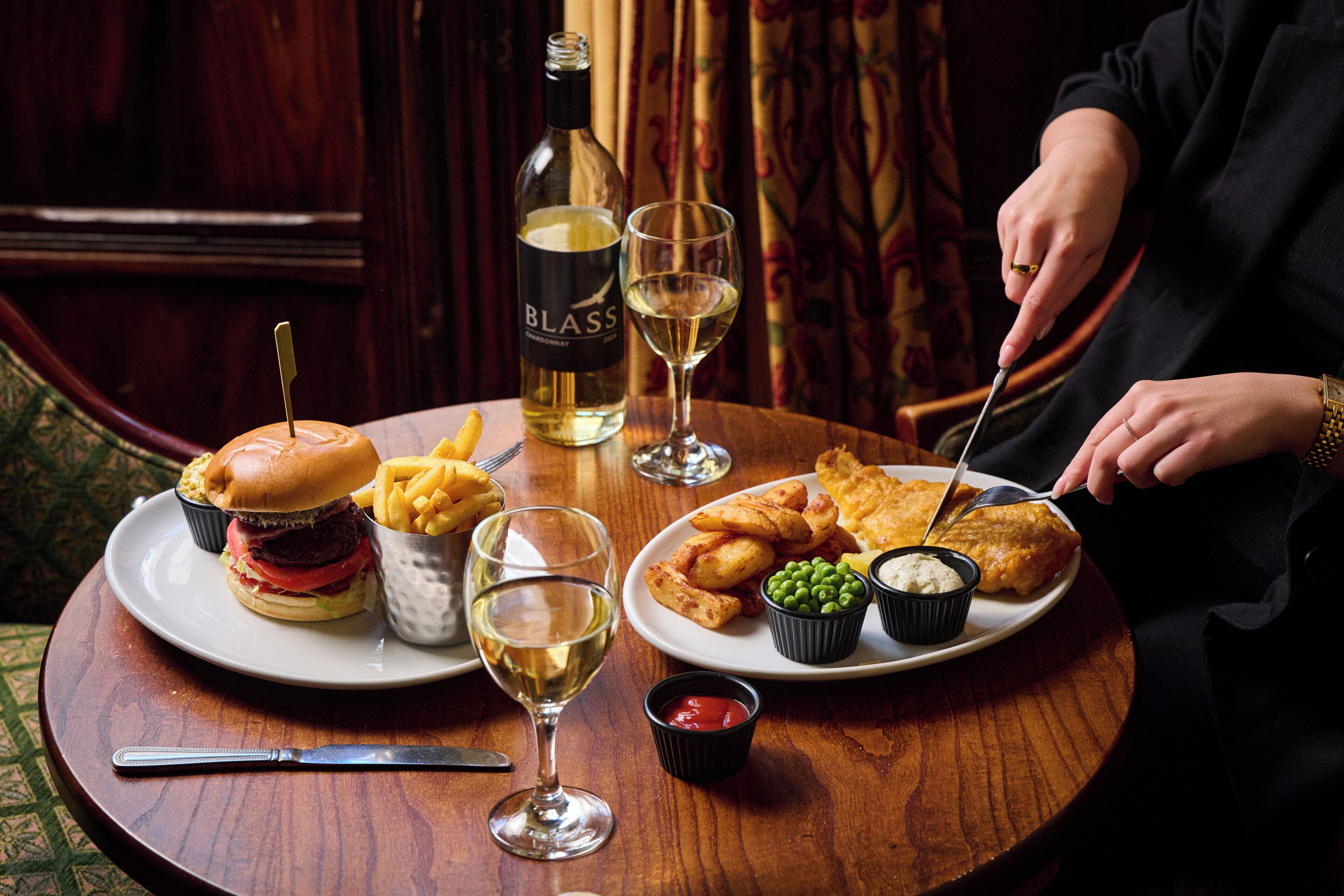 Une table à manger avec un hamburger, des frites, du poisson-frites et une bouteille de vin.