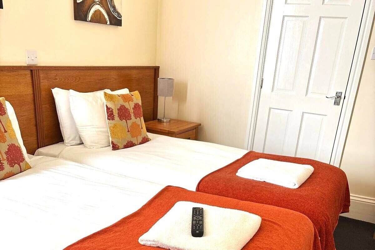 Habitación de hotel con dos camas, cada una con una toalla roja y un control remoto. Hay una mesita de noche con una lámpara, un cuadro en la pared y una puerta blanca.