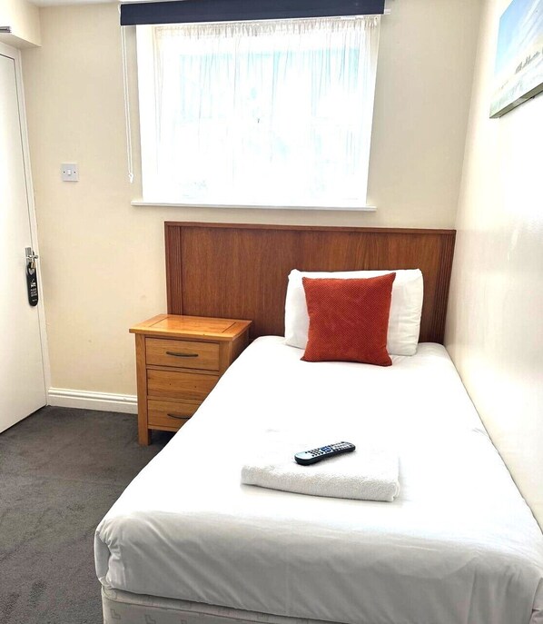 A hotel room with a bed, a wooden nightstand, a remote control, and a window with curtains.