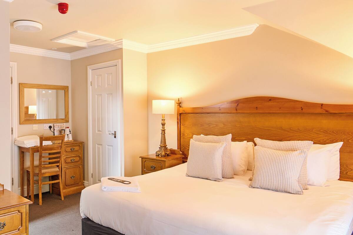 Habitación de hotel con una cama grande, cabecera de madera, escritorio con silla, espejo y mesita de noche con lámpara.