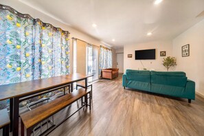 Een woonkamer met een houten eettafel, bankjes, een blauwgroene bank en een televisie aan de muur.