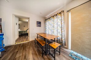 Een kamer met een houten eettafel en bankjes, een blauwe stoel, een gordijn met bloemenprint en een bord op de muur.