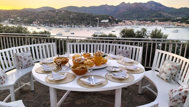 Mesa redonda dispuesta para una comida, con sillas blancas, con vista a un puerto con barcos y un telón de fondo montañoso.