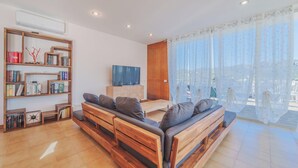 Una sala de estar moderna con un sofá de madera, una estantería, un televisor de pantalla plana y un amplio ventanal con cortinas translúcidas.