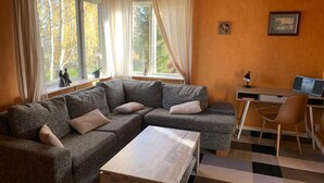 A cozy living room with a sectional sofa, a wooden coffee table, a desk with a computer and speakers, and large windows with curtains.