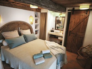 Una camera accogliente con un letto grande, una testiera in legno, una mensola sul comodino con oggetti decorativi, un bagno con lavandino e specchio, e una porta in legno rustica.