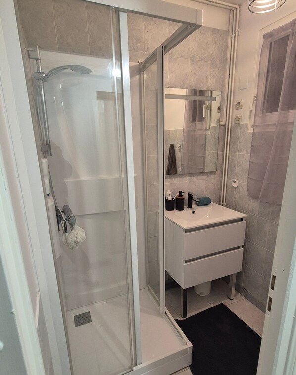Een moderne badkamer met een douchecabine van glas, een witte wastafel met spiegel, en een zwarte badmat.
