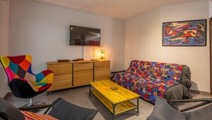 A modern living room with a colorful armchair, a sofa with a patterned cover, a wooden cabinet, and a flat-screen TV mounted on the wall. - Coeur d'Azur - apt climatisé - 1km plage (Saint Raphael)
