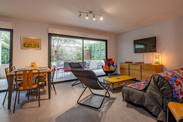 Een moderne woonkamer met een bank, een salontafel, een eettafel met stoelen, een kleurrijke fauteuil en een aan de muur bevestigde televisie.