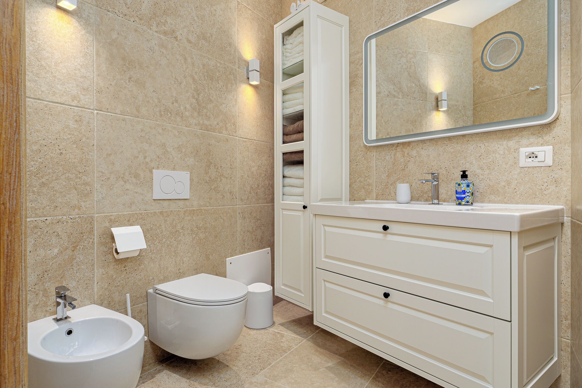 Une salle de bain comprenant des toilettes, un bidet, un meuble-lavabo avec un miroir et un espace pour ranger les serviettes.