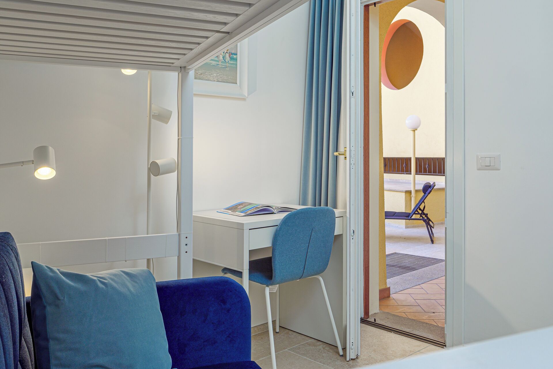 A modern room with a desk, a blue chair, and a view of an outdoor area with a lounge chair and a circular architectural feature.