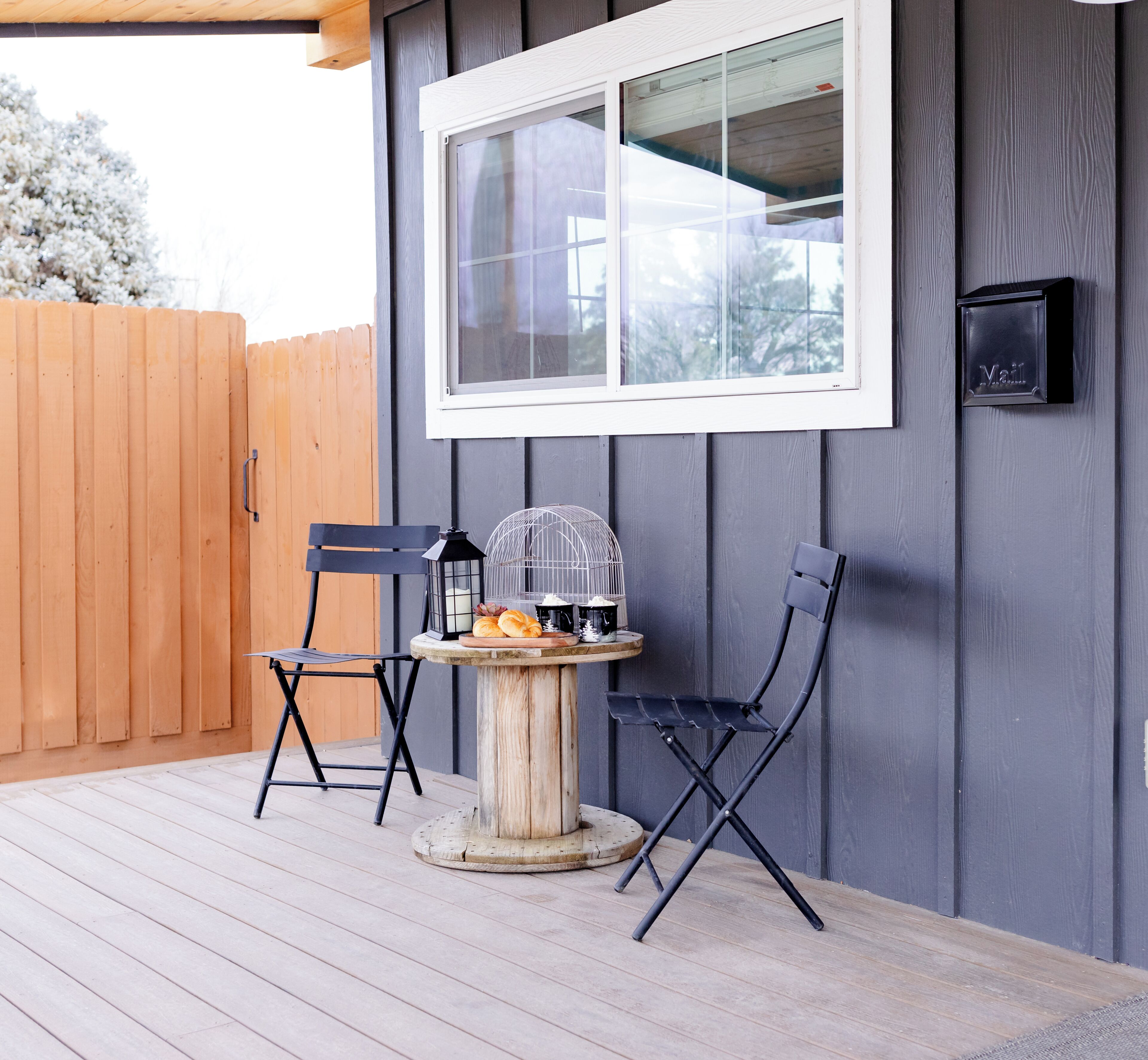 Une terrasse en bois avec deux chaises noires et une petite table ronde agrémentée d’une lanterne et d’une cage à oiseaux.