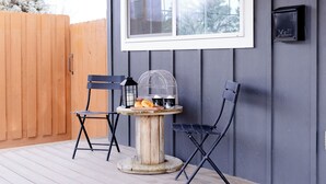 Una terraza de madera con dos sillas negras y una mesa redonda pequeña con un farol y una jaula de pájaros.