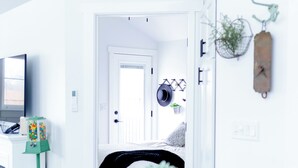 Una habitación moderna con decoración en blanco, que incluye un comedor con mesa y sillas blancas, una planta pequeña en maceta y una puerta que da a otra habitación.