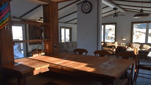 Un salon chaleureux avec une table à manger en bois, des sièges confortables, une horloge au mur et une vue sur un paysage enneigé par de grandes fenêtres.