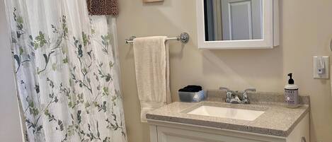 Un baño con cortina de ducha, toallero, lavabo con espejo y un pequeño cuadro en la pared.
