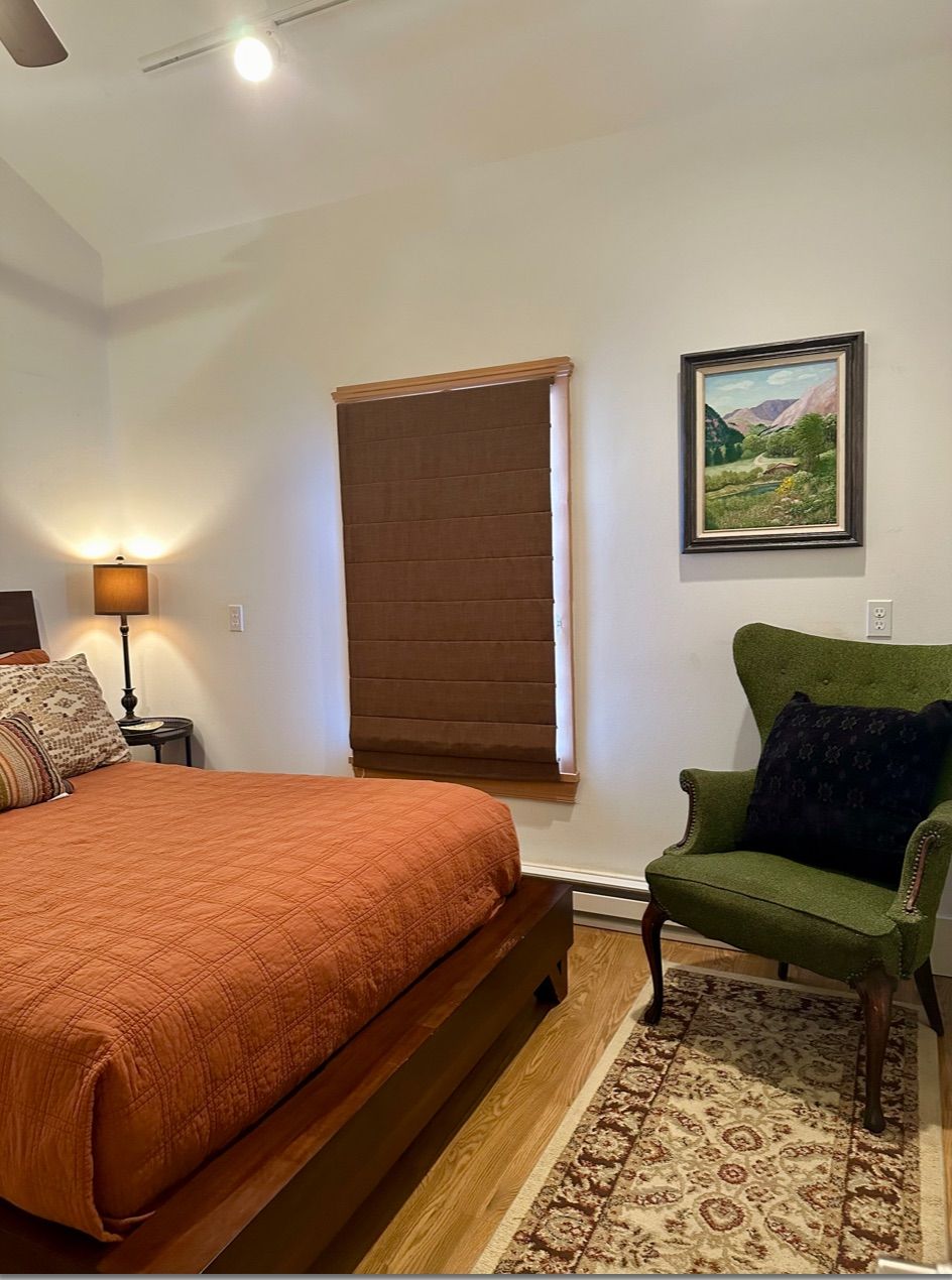 A bedroom with a bed, a chair, a window with blinds, a lamp, and a painting on the wall.