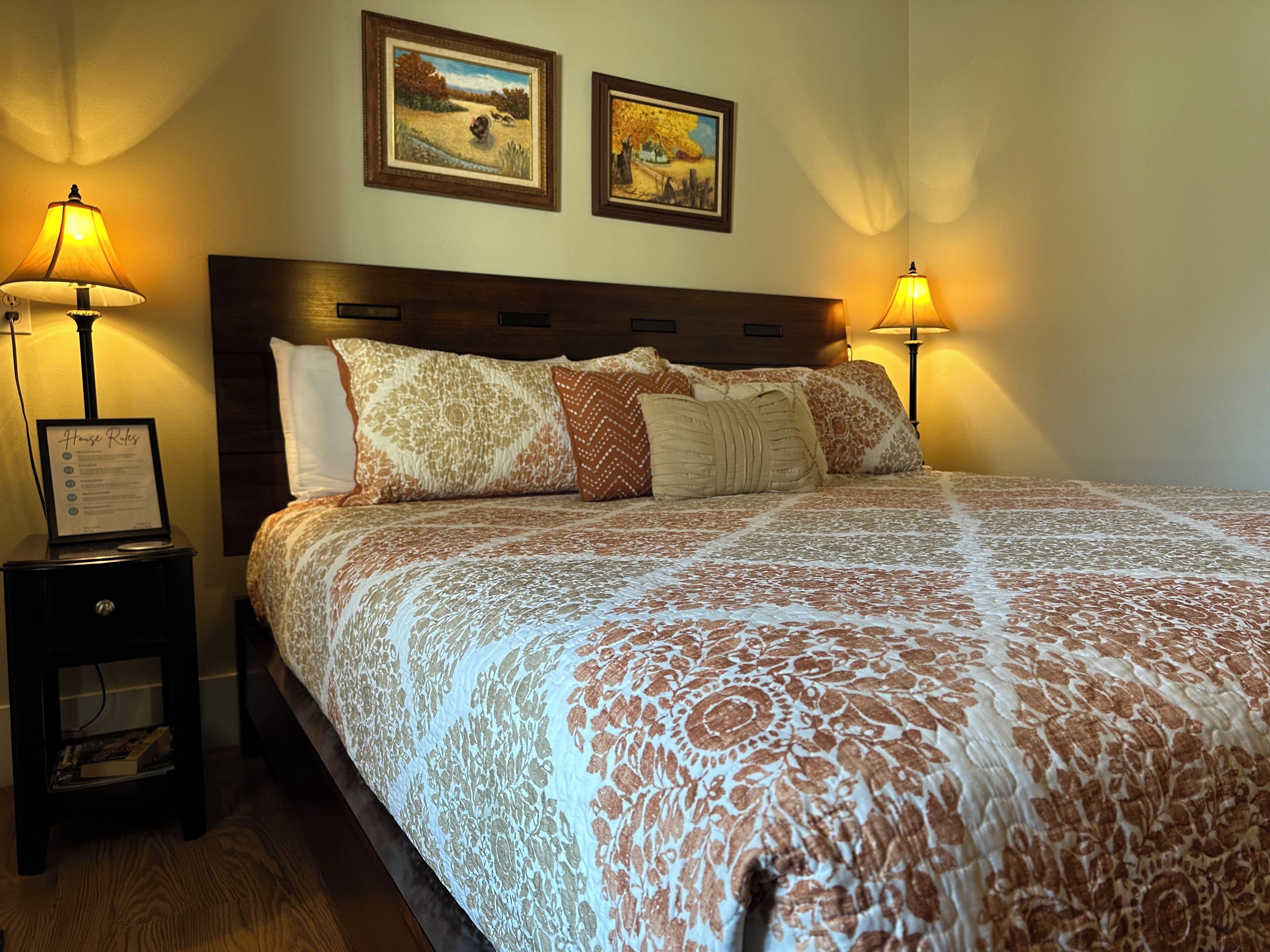 A neatly made bed with a patterned bedspread, two framed pictures on the wall, and two bedside lamps.