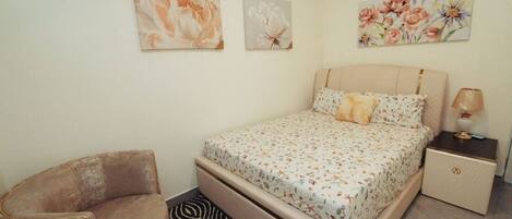Un dormitorio con cama, una silla, una mesita de noche y dos cuadros colgados en la pared.