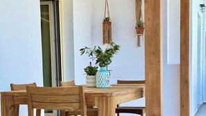 A wooden dining table with four chairs, a potted plant, and macrame wall hangings in a bright room with a tiled floor. - Famly Apartment with Sea View & Plunge Pool (Ground Floor) (Milos)