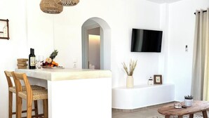 A modern kitchen with a central island, two wicker pendant lights, a flat-screen TV, and a dining area with a coffee table. - Famly Apartment with Sea View & Plunge Pool (Ground Floor) (Milos)