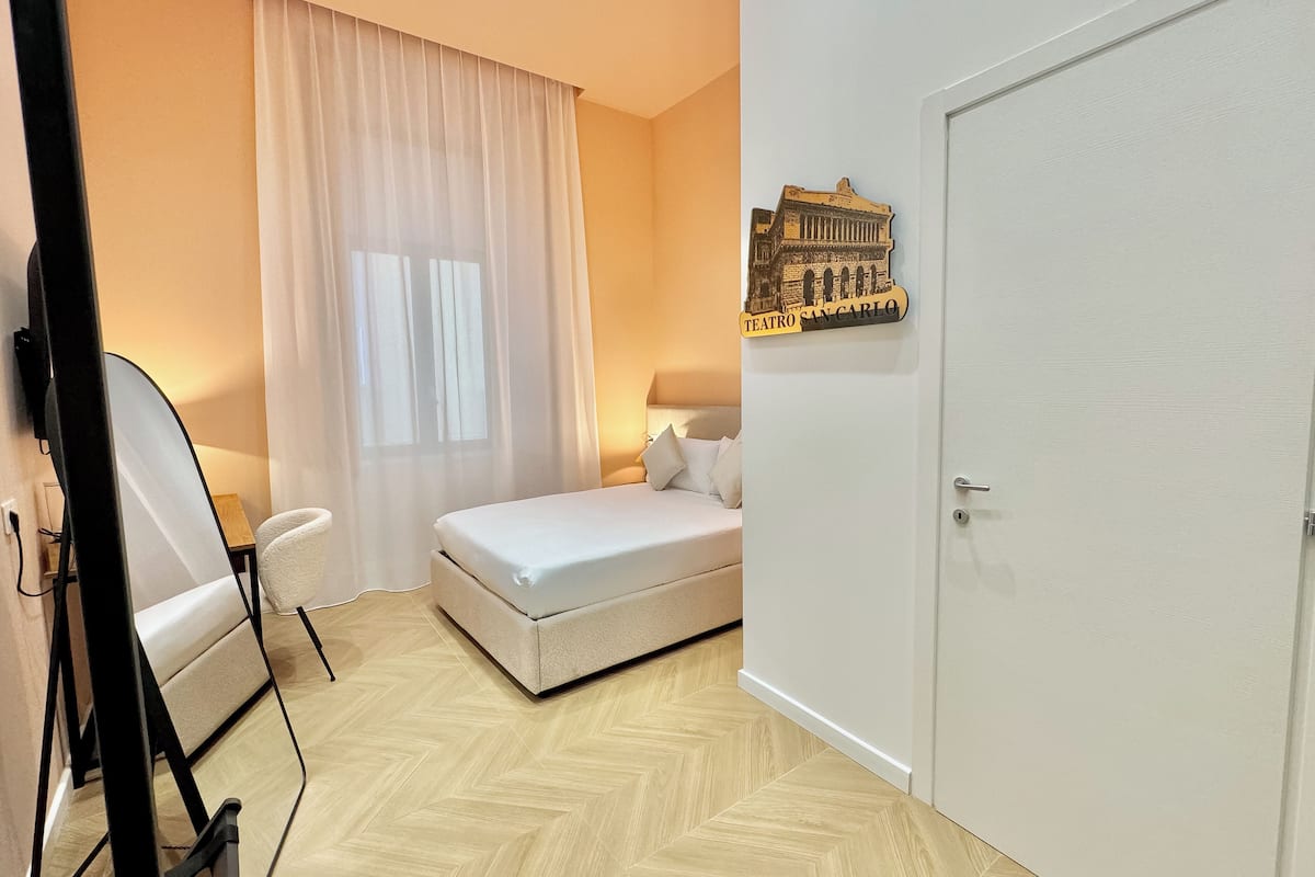 A hotel room with a bed, a mirror, a window with curtains, a wall-mounted picture, and a door.