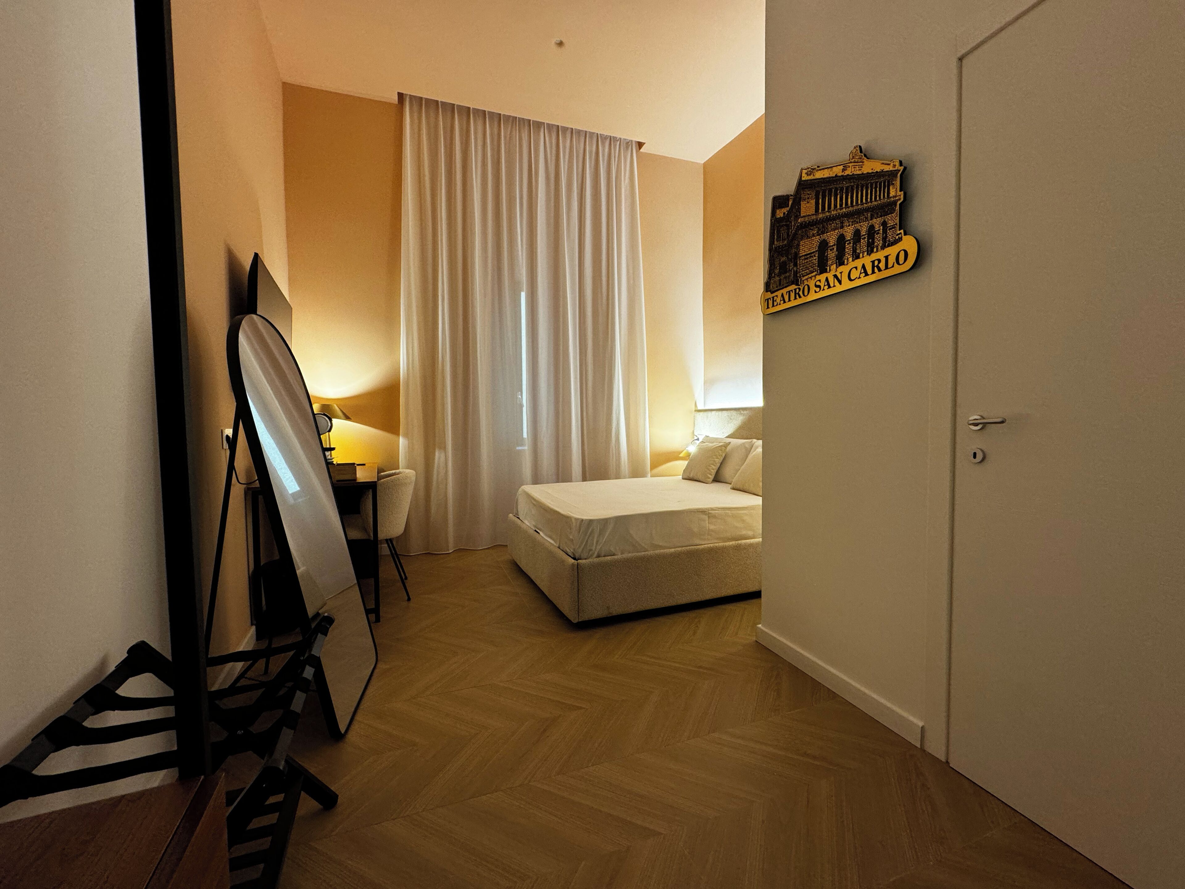 A hotel room with a bed, a desk with a lamp, a mirror, and a wall sign.
