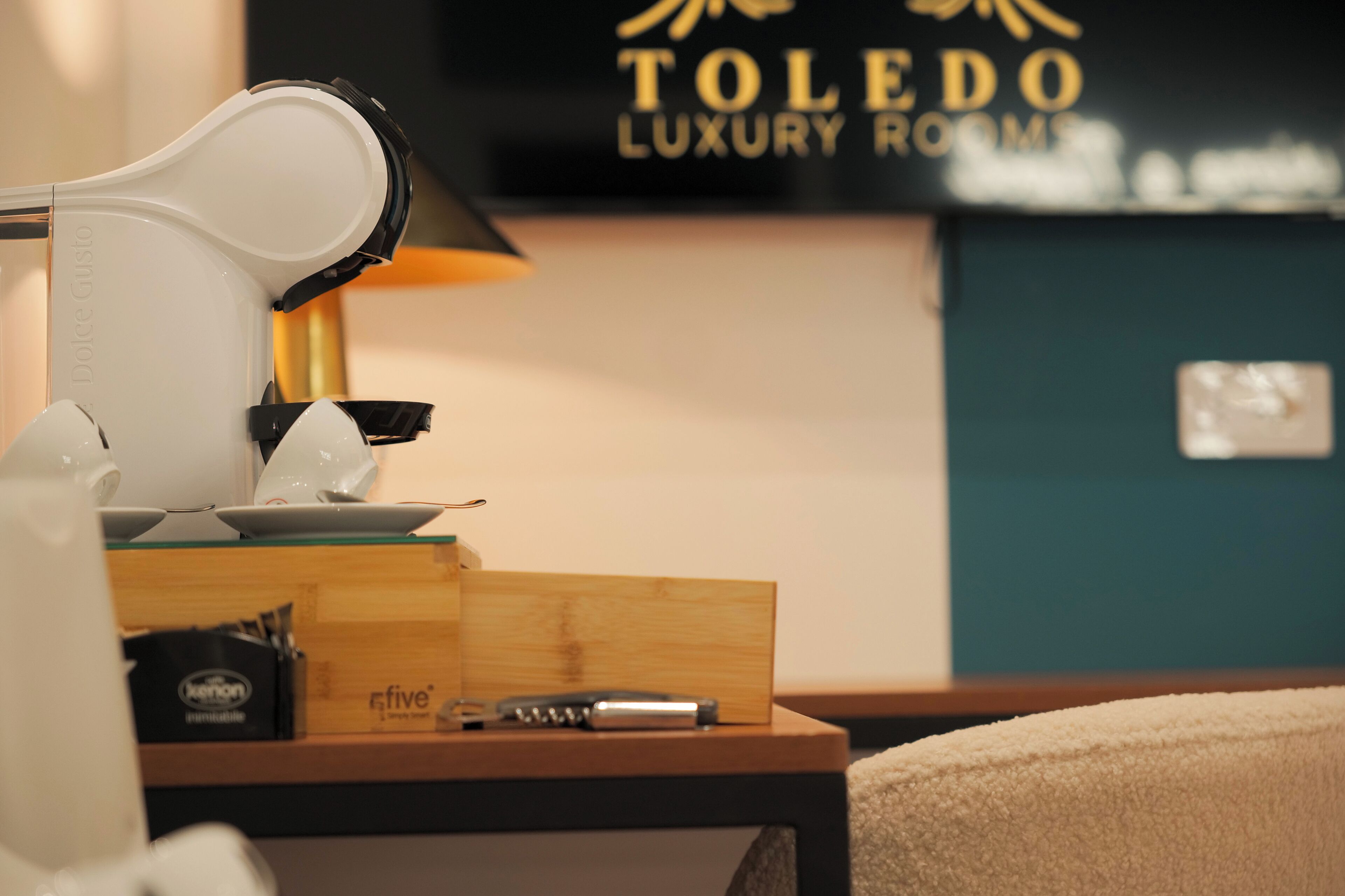Ein ordentlich eingerichtetes Hotelzimmer mit einer Dolce Gusto Kaffeemaschine, einem Stift und einem Smartphone auf einem Holznachttisch. Im Hintergrund ist ein Schild für Toledo Luxury Rooms zu sehen.